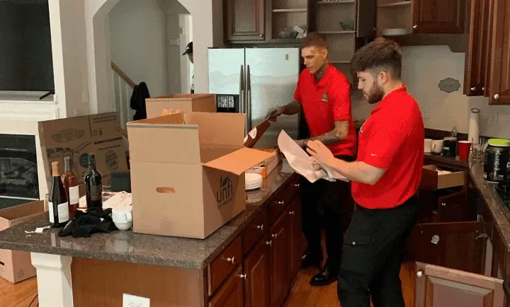 Two movers wrapping dishes with packing paper beside open kitchen boxes