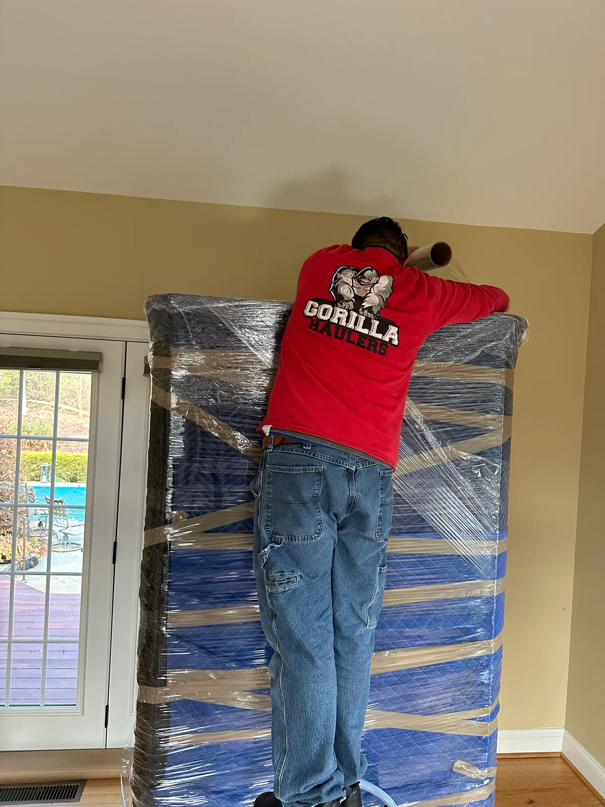 Gorilla Haulers mover stretch-wrapping a tall, blanket-padded armoire inside a house